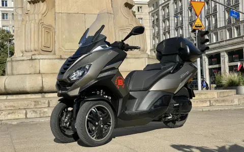Trójkołowy skuter Peugeot Metropolis 400 na tle zabytkowej kamienicy i pomnika.