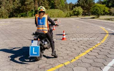Nauka jazdy motocyklem na placu manewrowym. Osoba w kamizelce odblaskowej z literą "L" na motocyklu.