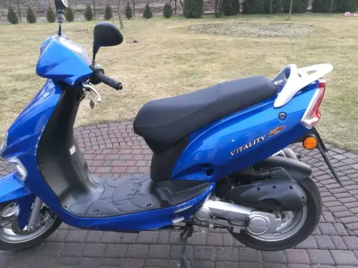 Błękitny skuter Kymco Vitality 50 2T stoi na brukowanym podjeździe, gotowy do jazdy.