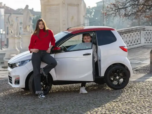 Młoda para przy białym samochodzie z czerwonym dachem. Ona opiera się o maskę, on siedzi za kierownicą. Idealni kandydaci na am1 prawo jazdy.