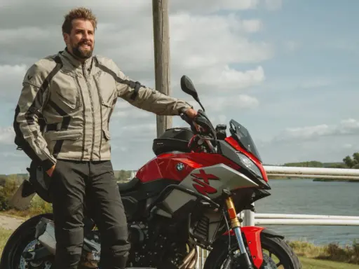 Uśmiechnięty motocyklista w kurtce od polskiego producenta odzieży motocyklowej, oparty o czerwoną maszynę nad jeziorem.