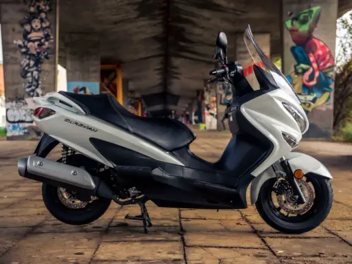 Biały skuter Suzuki Burgman 125, z czarnym siedzeniem, zaparkowany pod mostem z graffiti. Poznaj suzuki burgman 125 dane techniczne.
