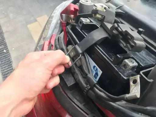 Ręka odłącza klemę akumulatora w skuterze. Akumulator znajduje się pod siedzeniem, obok tylnego światła.