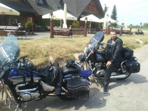 Motocyklista w skórzanej kurtce i spodniach odpoczywa przy swoim motocyklu. W tle karczma "Bida", idealne miejsce na postój podczas odkrywania **tras motocyklowych lubelskich**.