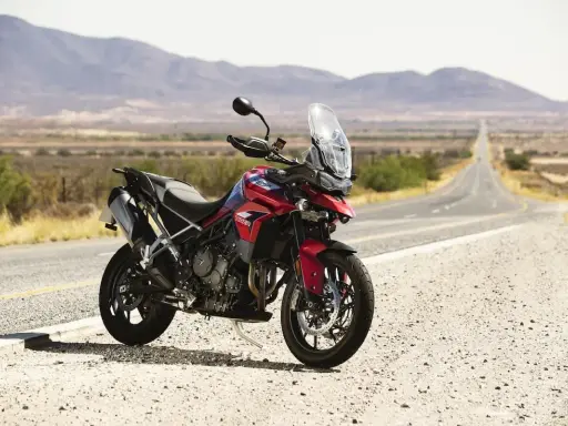 Czerwony motocykl Triumph Tiger 900 GT Pro zaparkowany na poboczu pustej drogi w krajobrazie z górami w tle.