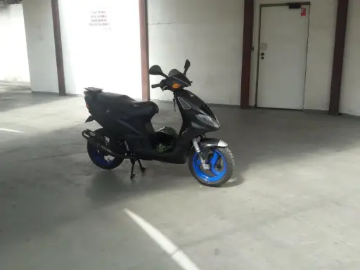 Czarny skuter Piaggio NRG MC3 z niebieskimi felgami stoi na betonowej posadzce w garażu. Obok niego leży zielony kask.