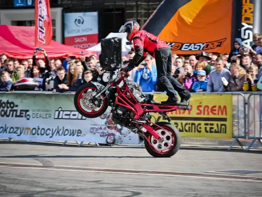 Motocykl do stuntu na tylnym kole, kierowca w czerwonej kurtce i kasku, tłum widzów na tle banerów.