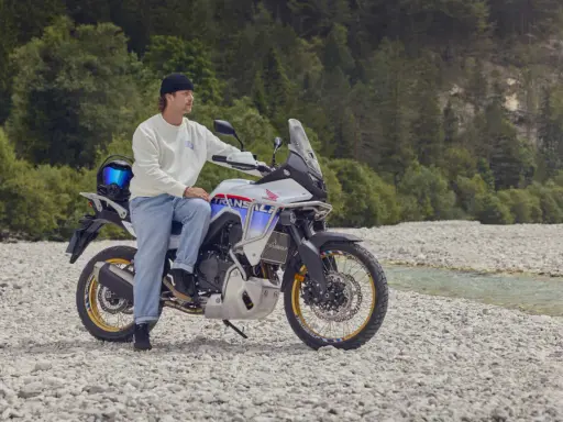 Mężczyzna na motocyklu Honda Transalp, gotowy na przygodę motocyklem po Europie. W tle las i strumień.