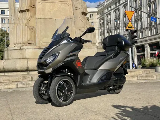 Trójkołowy skuter Peugeot Metropolis 400 na tle zabytkowej kamienicy i pomnika.