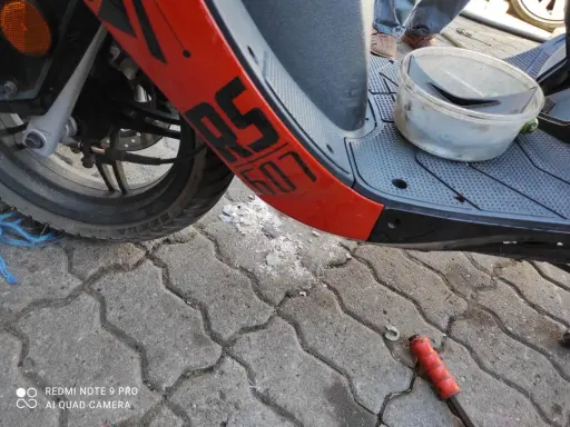 Część skutera Junak 607 4T z pomarańczowym elementem i napisem "607". Na podłodze leżą części i pojemnik.