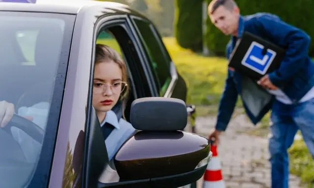 Dziewczyna za kierownicą auta szkoleniowego, instruktor z tablicą "L" obok. Spełnienie wymagań na prawo jazdy kat. A.