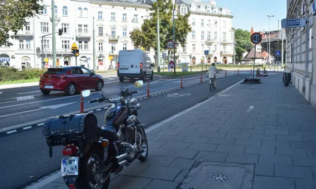 Motocykl zaparkowany na chodniku, obok drogi z wyznaczonym pasem rowerowym. W tle budynki i ruch uliczny, w tym czerwony samochód i biały van.