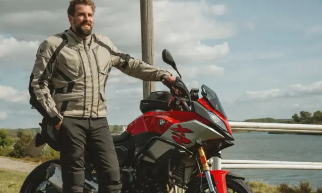 Uśmiechnięty motocyklista w kurtce od polskiego producenta odzieży motocyklowej, oparty o czerwoną maszynę nad jeziorem.
