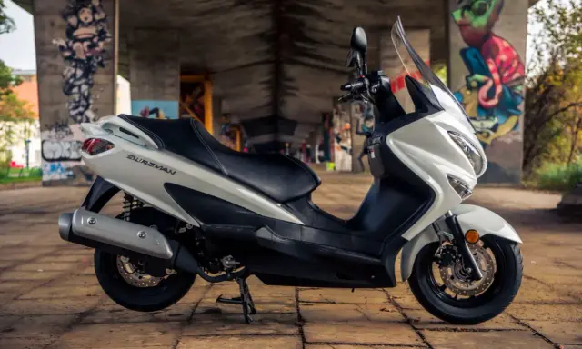 Biały skuter Suzuki Burgman 125, z czarnym siedzeniem, zaparkowany pod mostem z graffiti. Poznaj suzuki burgman 125 dane techniczne.