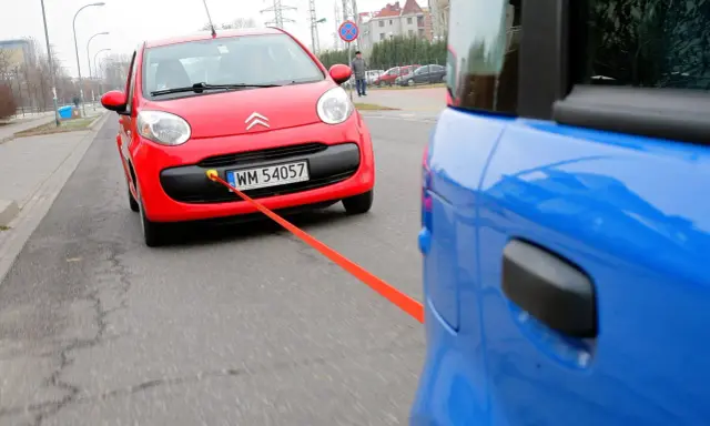 Czerwony Citroen holowany przez niebieski samochód. Pomarańczowa linka holownicza jest napięta, sugerując ruch i prędkość.