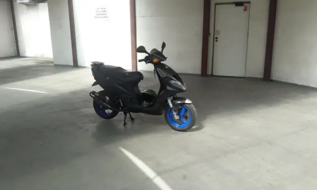 Czarny skuter Piaggio NRG MC3 z niebieskimi felgami stoi na betonowej posadzce w garażu. Obok niego leży zielony kask.