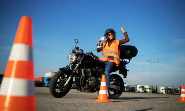 Szczęśliwa motocyklistka z prawem jazdy kategorii A3 w ręku, siedząca na motocyklu, świętuje sukces na placu manewrowym.