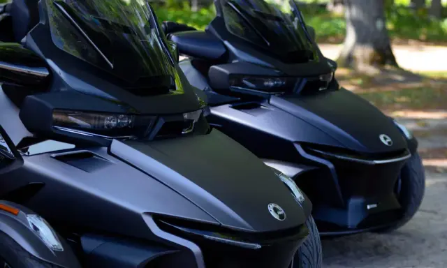 Dwa matowe, czarne pojazdy typu Can-Am Spyder stoją obok siebie na parkingu dla motocykli, gotowe do jazdy.