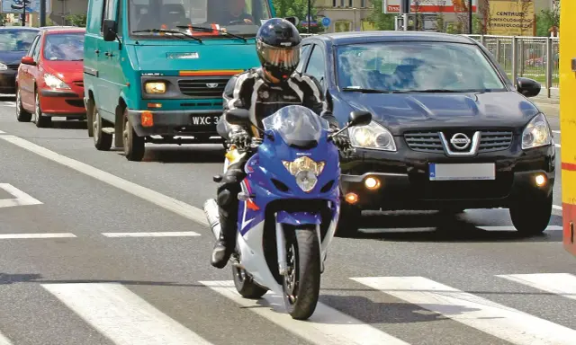 Motocyklista w kasku i kombinezonie na pasach. Obok samochody i furgonetka. Pamiętaj o przeglądzie, bo jaka kara za brak przeglądu może być dotkliwa.