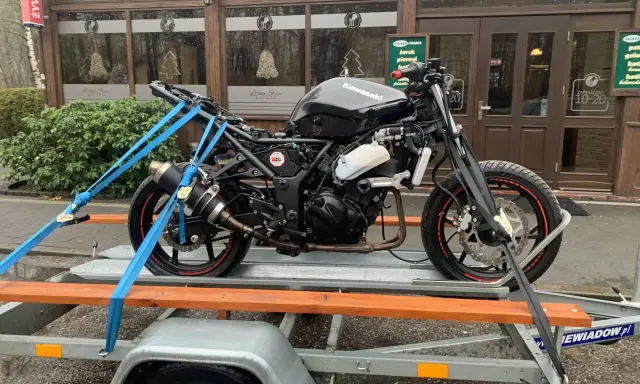 Czarny motocykl Kawasaki na przyczepce, zabezpieczony pasami. W tle budynek z napisem "Leśny Staw". Przepisy dotyczące przyczepki do motocykla są kluczowe dla bezpiecznego transportu.