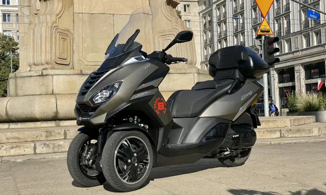Trójkołowy skuter Peugeot Metropolis 400 na tle zabytkowej kamienicy i pomnika.