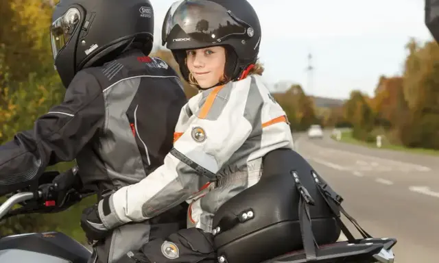 Uśmiechnięte dziecko na motocyklu, w kasku i kurtce, siedzi za kierowcą.