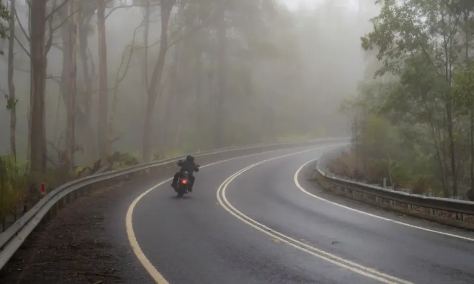 Motocyklista pokonuje zakręt na mokrej drodze wśród mglistego lasu. Jazda motocyklem w deszczu wymaga skupienia.