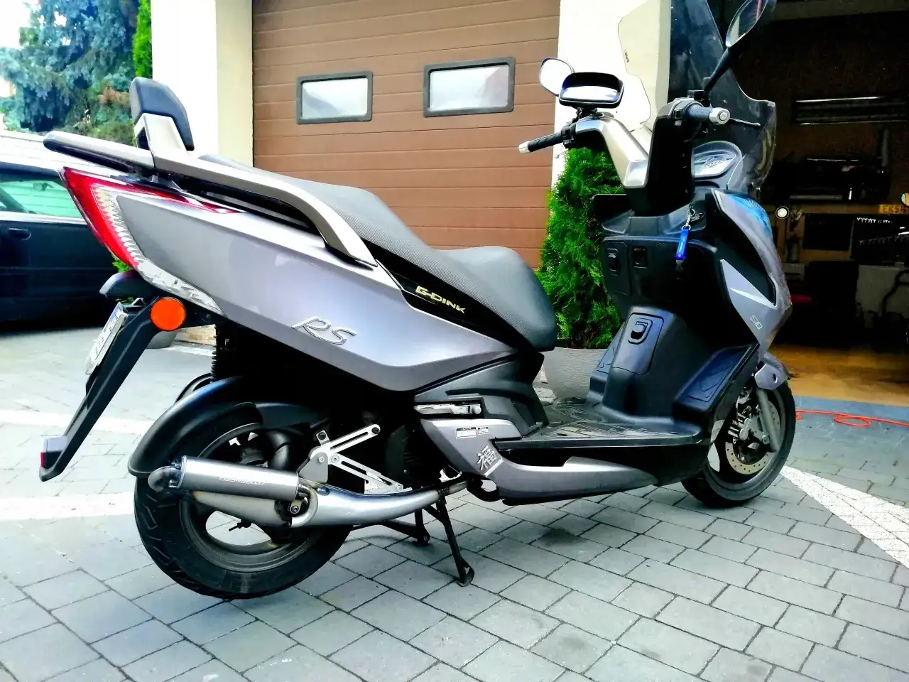 Szary skuter Kymco Dink 50 z czarnym siedzeniem i sportowym wydechem Giannelli stoi na kostce brukowej przed garażem.