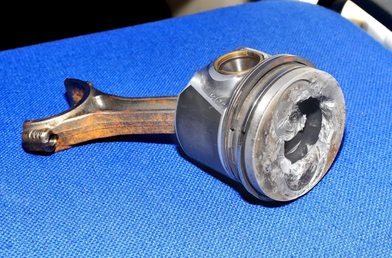 Pęknięty tłok objawy: widoczny poważnie uszkodzony tłok z korbowodem, metaliczne odłamki i głębokie pęknięcia.