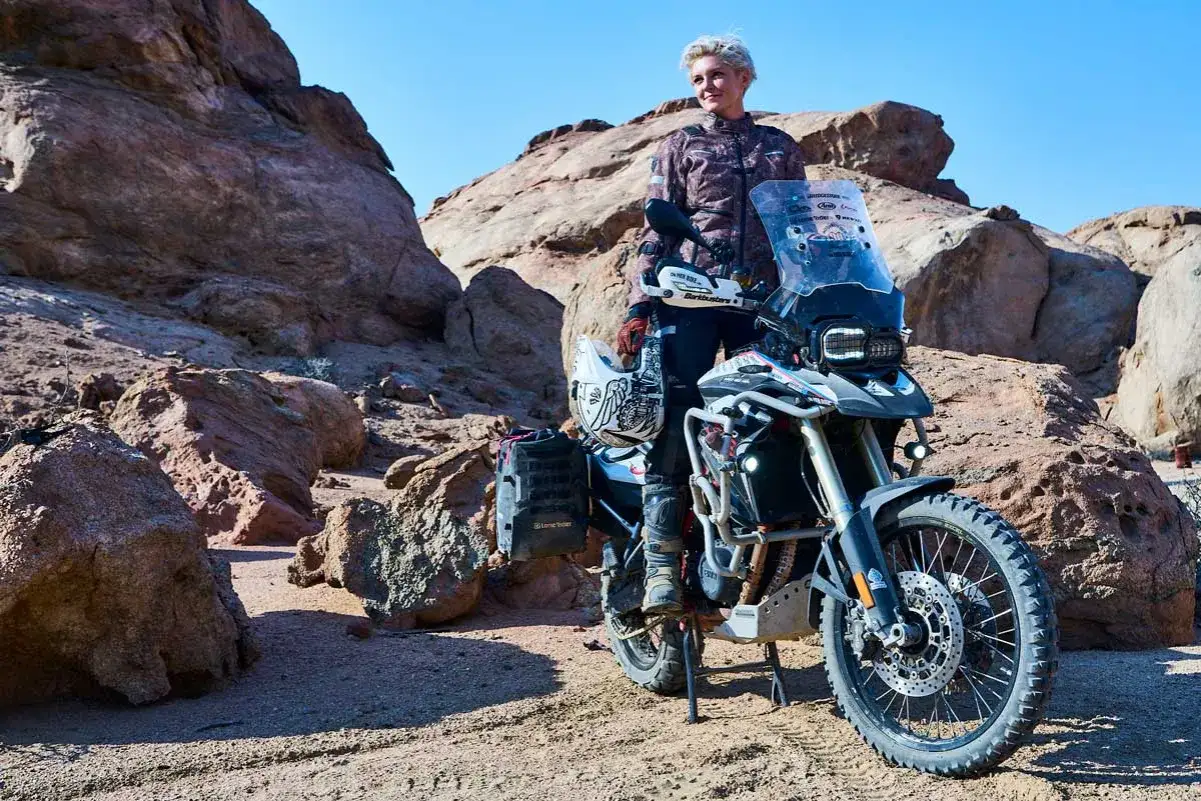 Kobieta w stroju motocyklowym stoi obok swojego motocykla w pustynnym krajobrazie. Jaki przebieg dla motocykla jest duży, gdy przemierza takie tereny?