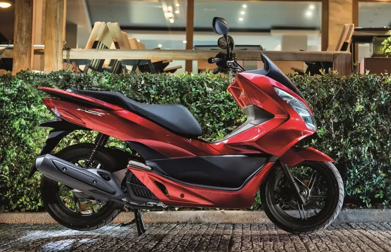 Czerwony skuter retro 125, Honda PCX, zaparkowany przed żywopłotem i restauracją.