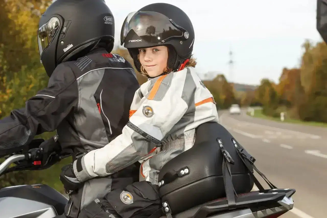 Uśmiechnięte dziecko na motocyklu, w kasku i kurtce, siedzi za kierowcą.