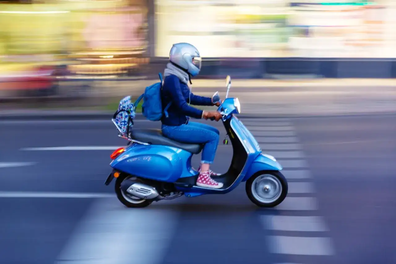Kobieta w kasku jedzie niebieskim skuterem 125. Czy skuterem 125 można jeździć po autostradzie? To pytanie nurtuje wielu kierowców.