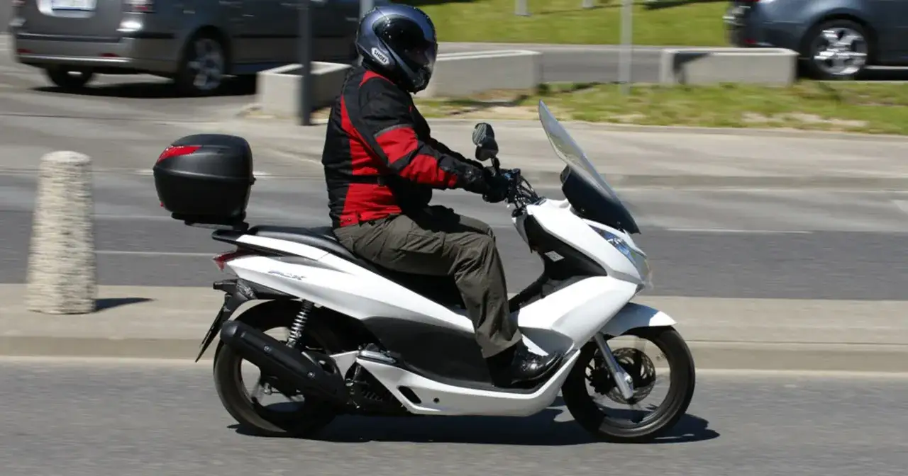 Motocyklista w kasku i kurtce jedzie białym skuterem. Czy skuterem 125 można jeździć po autostradzie?