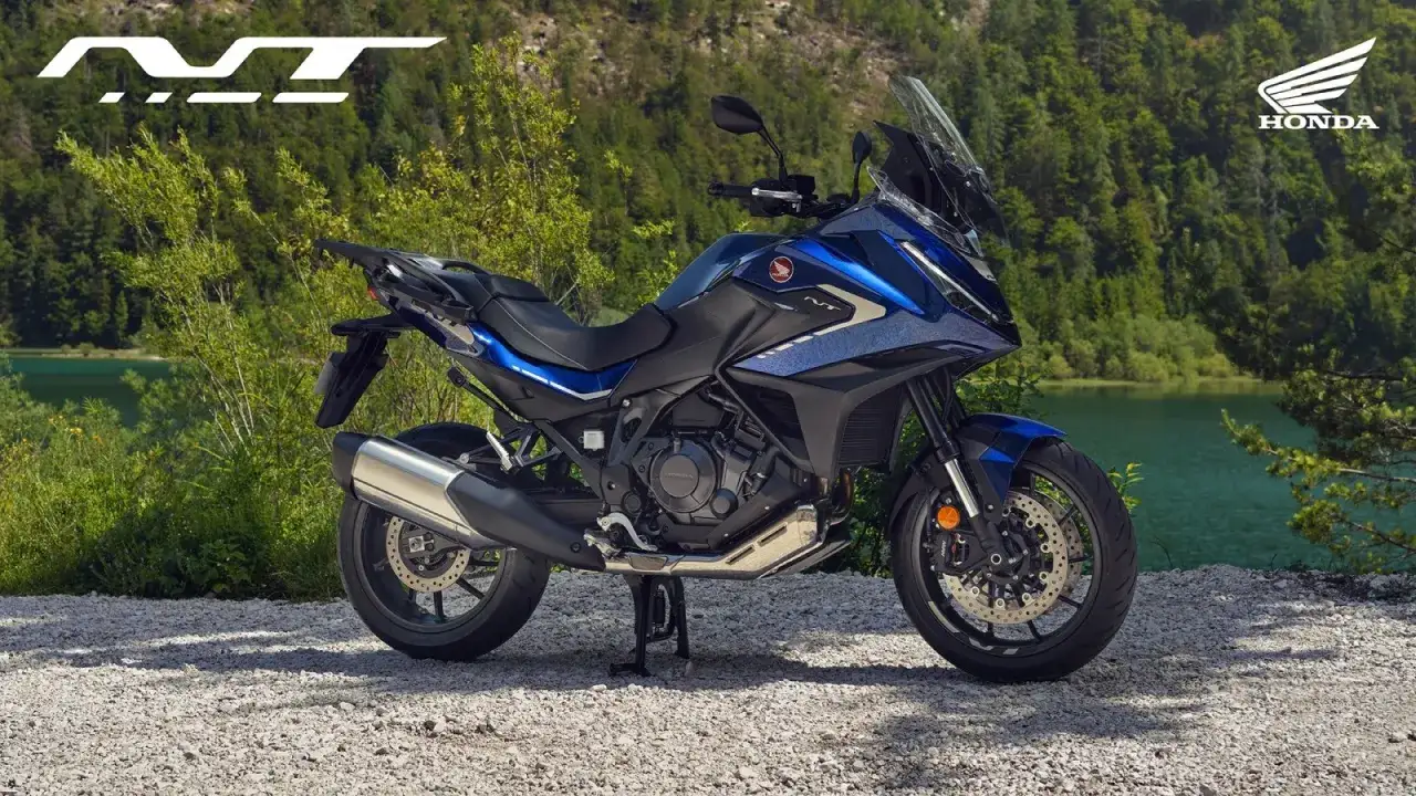 Niebieski motocykl Honda NT 1100 zaparkowany na żwirowej drodze, z jeziorem i lasem w tle.