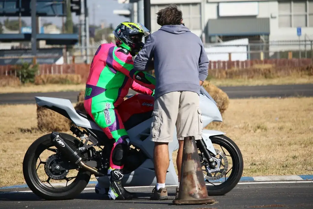 Kierowca w kolorowym kombinezonie przygotowuje się do jazdy na motocyklu, a instruktor udziela mu ostatnich wskazówek.