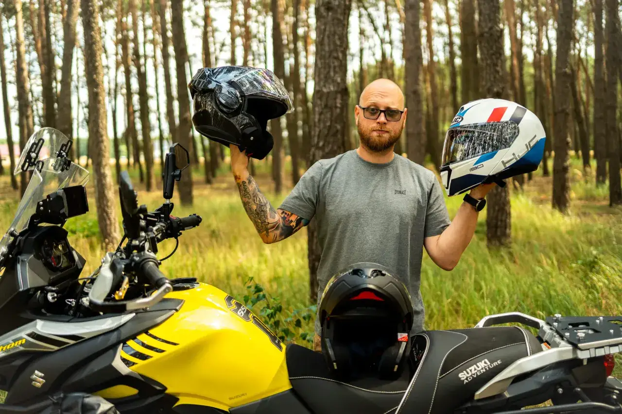 Mężczyzna z tatuażami porównuje kask szczękowy i integralny, stojąc obok żółtego motocykla Suzuki w lesie.
