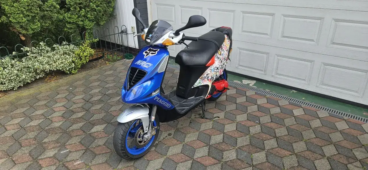 Niebieski skuter Piaggio NRG MC3 z naklejkami, zaparkowany na podjeździe przed garażem.