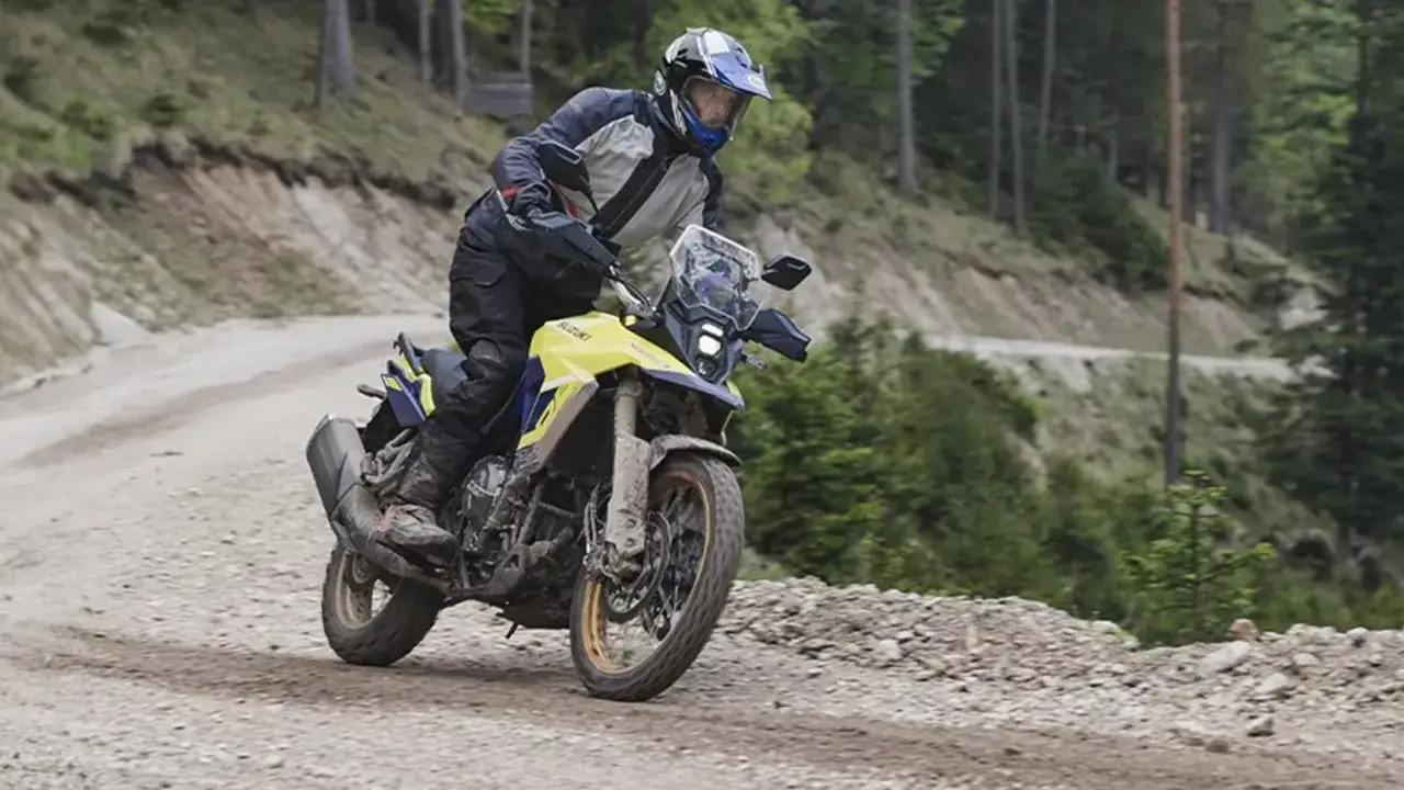Motocyklista w kasku i stroju terenowym pokonuje kamienistą drogę na żółto-niebieskim motocyklu Suzuki V-Strom 800 DE, otoczony zielonym lasem.
