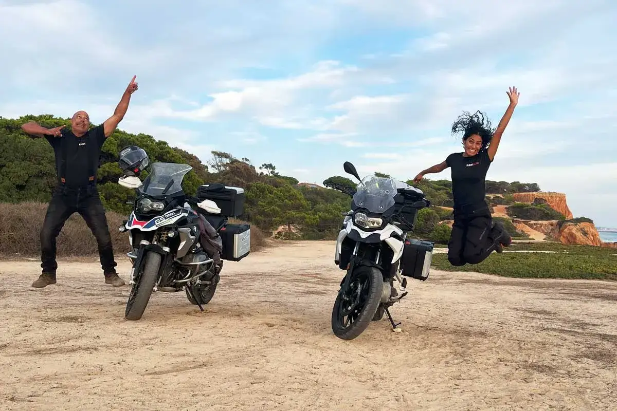 Dwóch motocyklistów, każdy z motocyklem 500 cm3, skacze z radości na tle malowniczego krajobrazu.