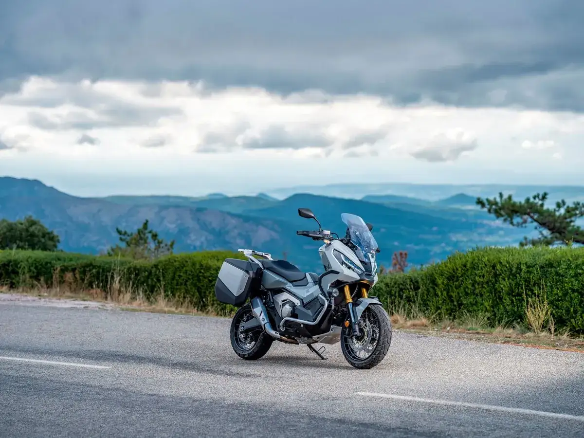 Szary motocykl Honda X-ADV 750 z kuframi stoi na drodze z widokiem na górzysty krajobraz.
