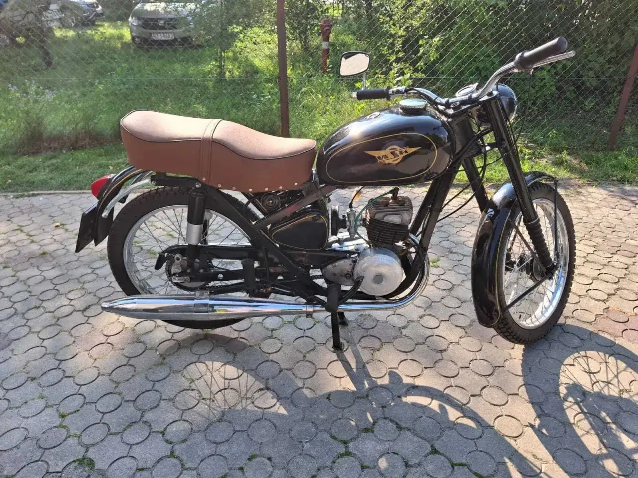 Klasyczny motocykl WSK 125 w kolorze czarnym, z brązowym siedzeniem, zaparkowany na chodniku z kostki brukowej.