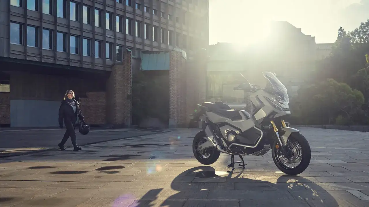 Kobieta w stroju motocyklowym idzie obok swojego motocykla Honda X-ADV 750, który stoi na parkingu przed nowoczesnym budynkiem.