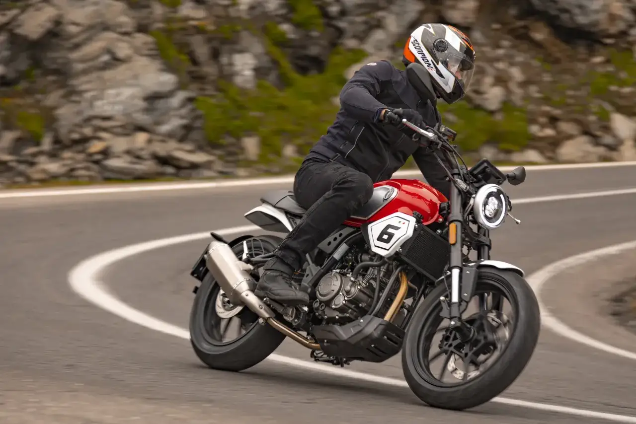 Motocyklista w kasku pokonuje zakręt na górskiej drodze. Czerwony motocykl 125ccm jest w trakcie docierania, a jego silnik pracuje płynnie.