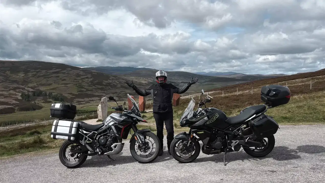 Motocyklista w kasku i kurtce pozuje obok dwóch motocykli: szarego i czarnego Tiger 900 GT Pro. W tle malowniczy krajobraz górski.