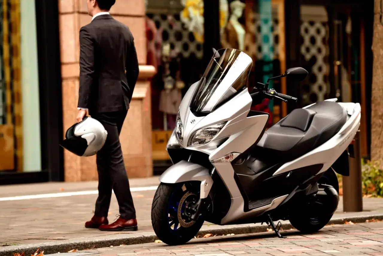 Elegancki mężczyzna z kaskiem obok srebrnego Suzuki Burgman 400. Wady i zalety tego skutera to temat dyskusji.