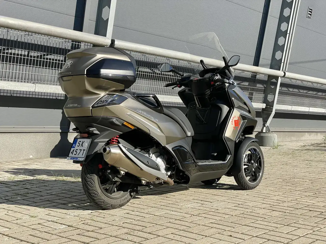 Złoty Peugeot Metropolis 400 z kufrem, zaparkowany na kostce brukowej przed nowoczesnym budynkiem.