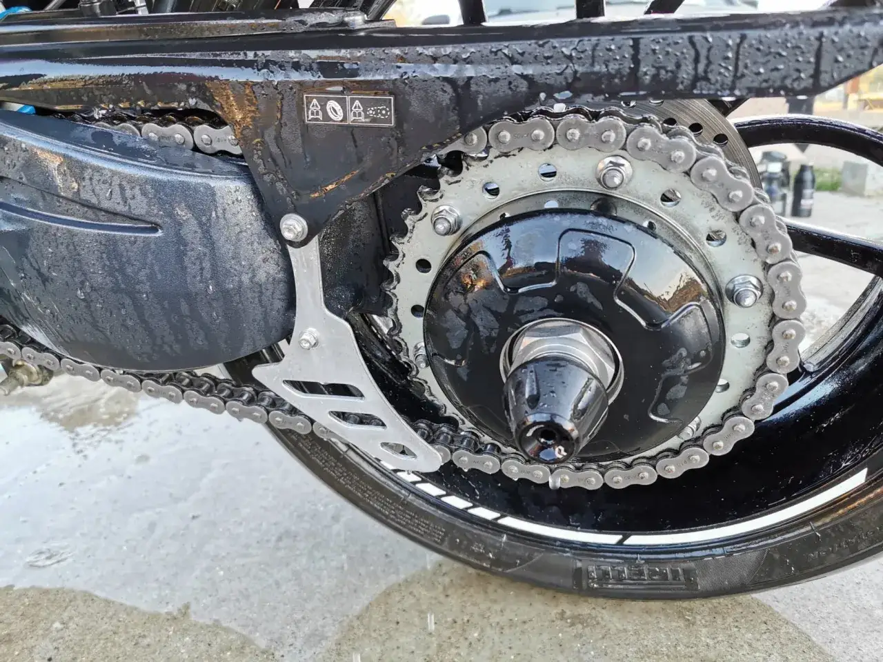 Motocykl z mokrym łańcuchem i zębatką. Widać elementy układu napędowego, sugerujące automatyczne smarowanie łańcucha.