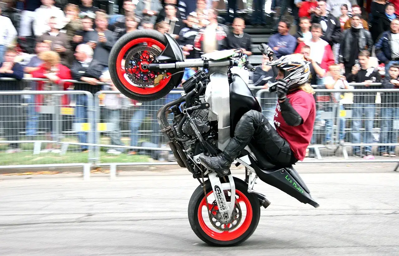 Motocykl do stuntu na tylnym kole, kierowca w kasku i kombinezonie, tłum widzów za barierkami.