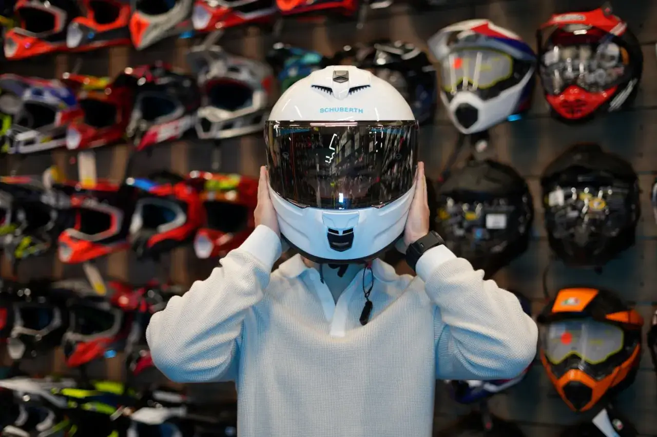 Osoba przymierza biały kask Schuberth. Czy to kask szczękowy czy integralny? W tle widać wiele innych kasków motocyklowych.